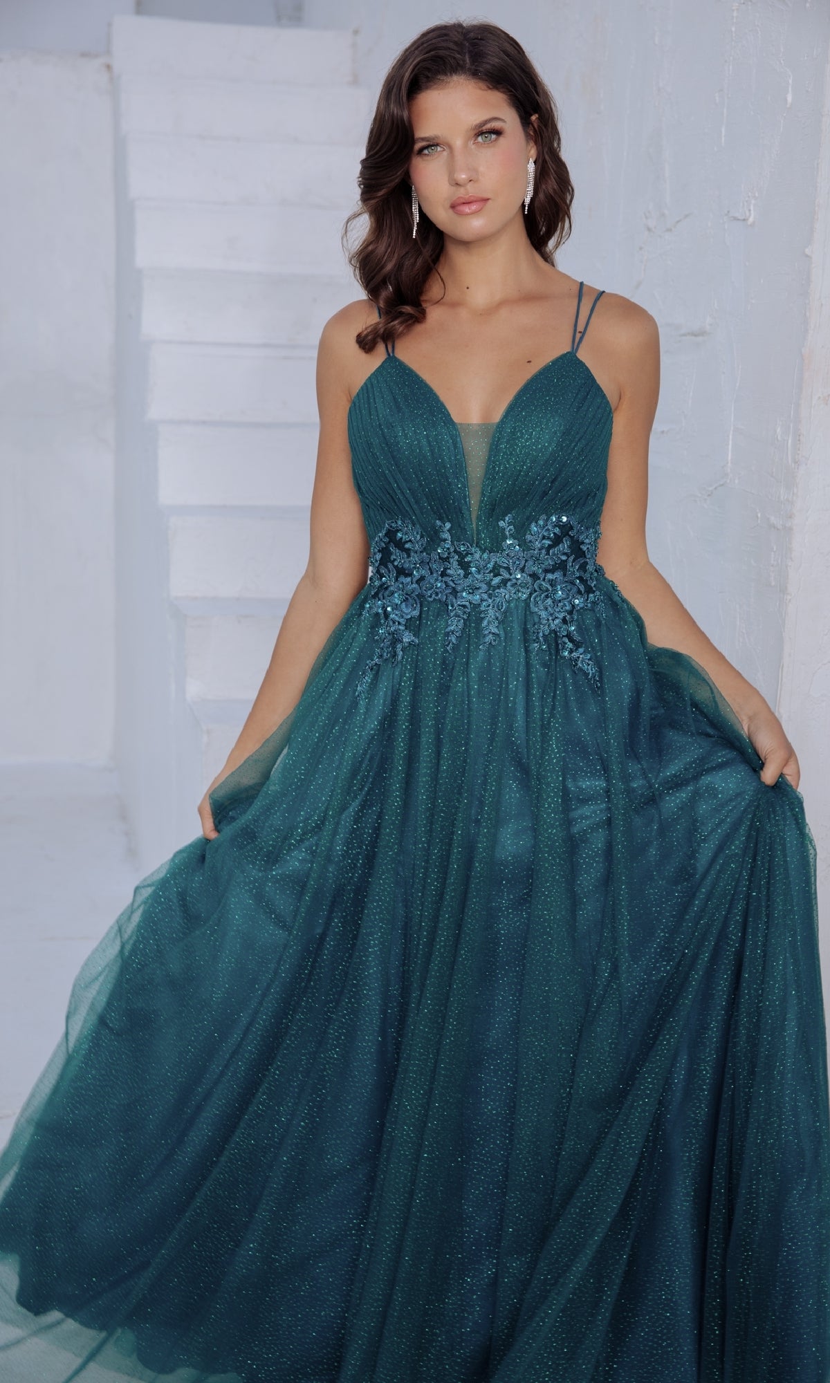 A woman in the Eureka 9797 Lace-Up Long Shimmer Prom Dress, a teal, floor-length A-line gown with thin straps and floral bodice details, stands before white stairs and walls.